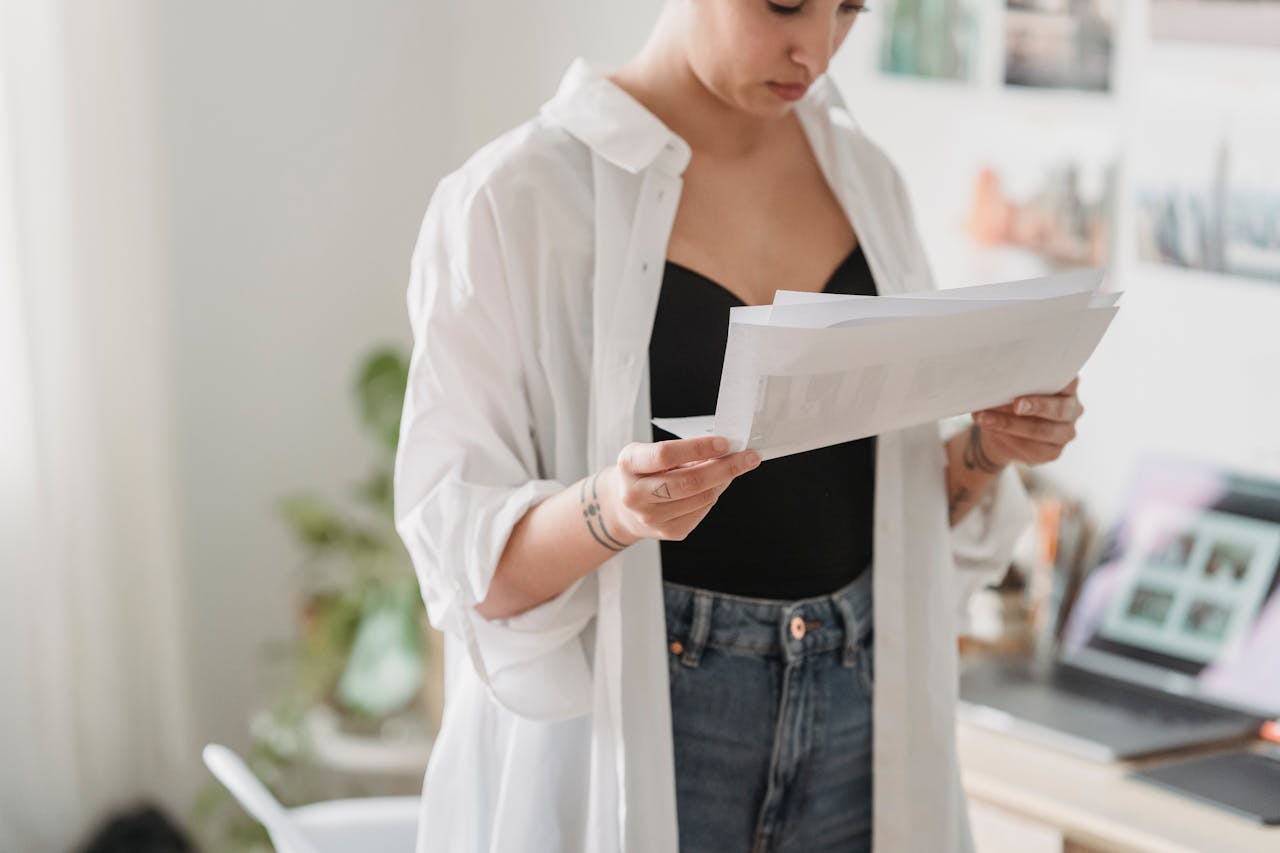 Crop young thoughtful woman with tattoos and paper sheets working against laptop at home in daytime